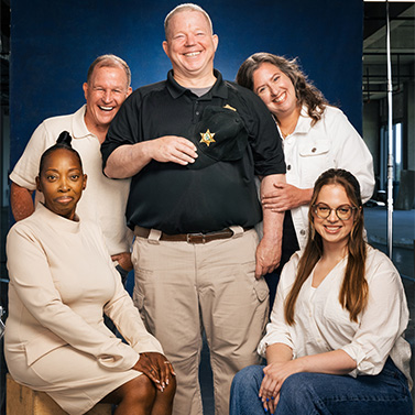 Five adults pose together, smiling warmly. One man in the center wears a black shirt with a sheriff’s badge and tan pants. Two women sit in front, while another man and woman stand behind, all against a dark blue background.