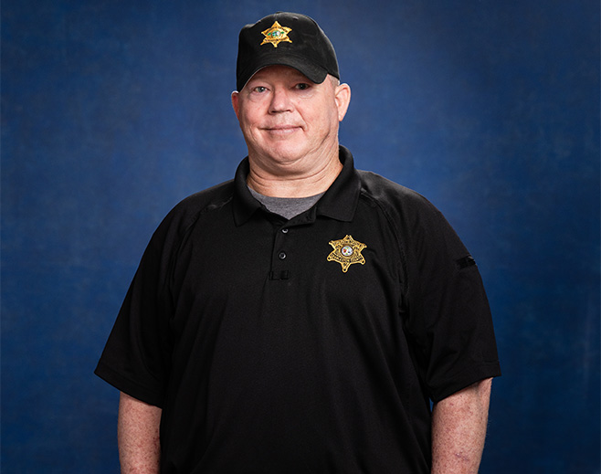 A man wearing a black cap and black polo shirt with a sheriff's badge emblem stands in front of a blue background, facing the camera and smiling slightly.