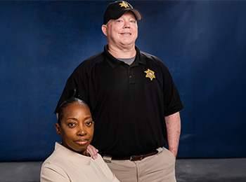 Two law enforcement officers pose for a portrait. One is a woman seated in a light-colored suit, and the other is a man standing behind her, wearing a black uniform and cap with a sheriff's badge. Blue background.