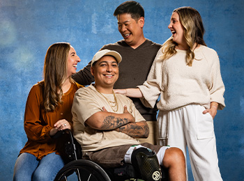 Four adults smile and laugh together against a blue background. One person sits in a wheelchair, while the other three stand close by, showing warmth and connection.
