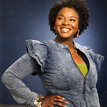 A woman with curly hair smiles confidently, standing with one hand on her hip. She wears a ruffled denim jacket over a green top, accessorized with a necklace and a bracelet, against a blue studio background.