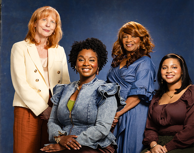 Four women pose together in front of a blue backdrop, smiling. One stands on the left in a white blazer; the other three are seated or standing, wearing blue and brown outfits. They appear relaxed and confident.