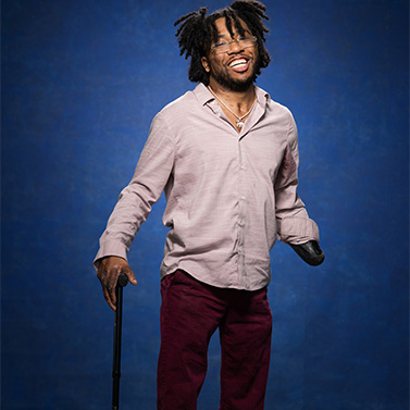 A smiling man with natural hair stands against a blue background, wearing a light pink button-up shirt and burgundy pants. He has a prosthetic arm and uses a cane for support.