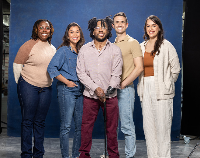 Five people stand together smiling in front of a blue backdrop. They are dressed casually and appear friendly and relaxed. One person in the center uses a prosthetic arm and holds a cane.