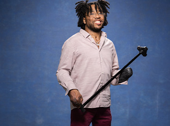 A smiling man with short dreadlocks stands against a blue background, wearing glasses, a light pink shirt, and maroon pants. He holds a cane in one hand and has a black prosthetic on the other arm.
