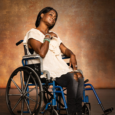 A woman in a wheelchair poses confidently against a warm, textured background. She wears a white top and ripped jeans, with one hand on her chest and the other resting on the chair’s armrest.