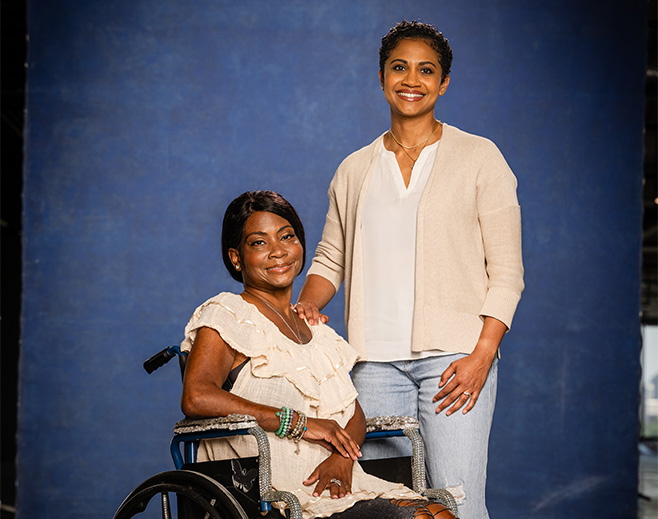 Two women pose together in front of a blue backdrop, one seated in a wheelchair wearing a cream dress, and the other standing beside her in a cardigan and jeans, both smiling warmly at the camera.