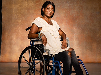 A woman in a wheelchair smiles at the camera. She is wearing a light-colored blouse and black pants, with her hair styled neatly and bracelets on her wrist, sitting against a warm, textured background.