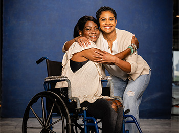 Two women smiling and hugging; one is seated in a wheelchair while the other kneels beside her. They are posed in front of a blue background.