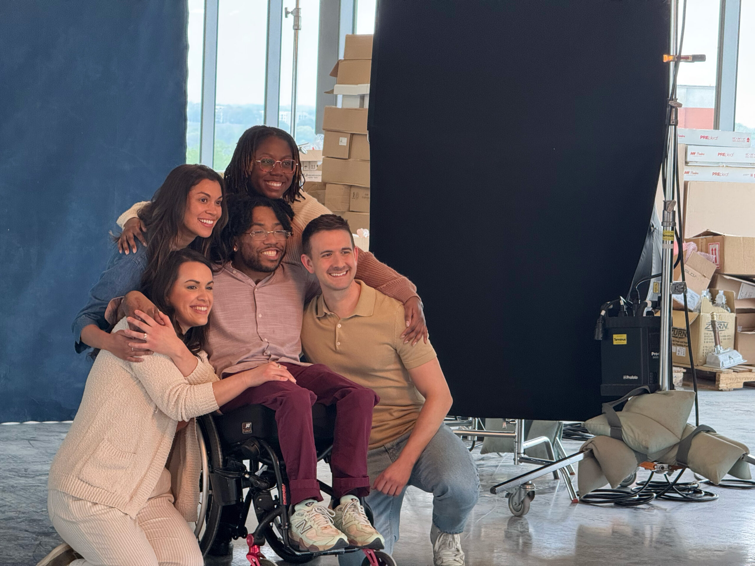 Five people, including a man in a wheelchair, pose closely together and smile for a photo in a bright studio with boxes and equipment in the background.