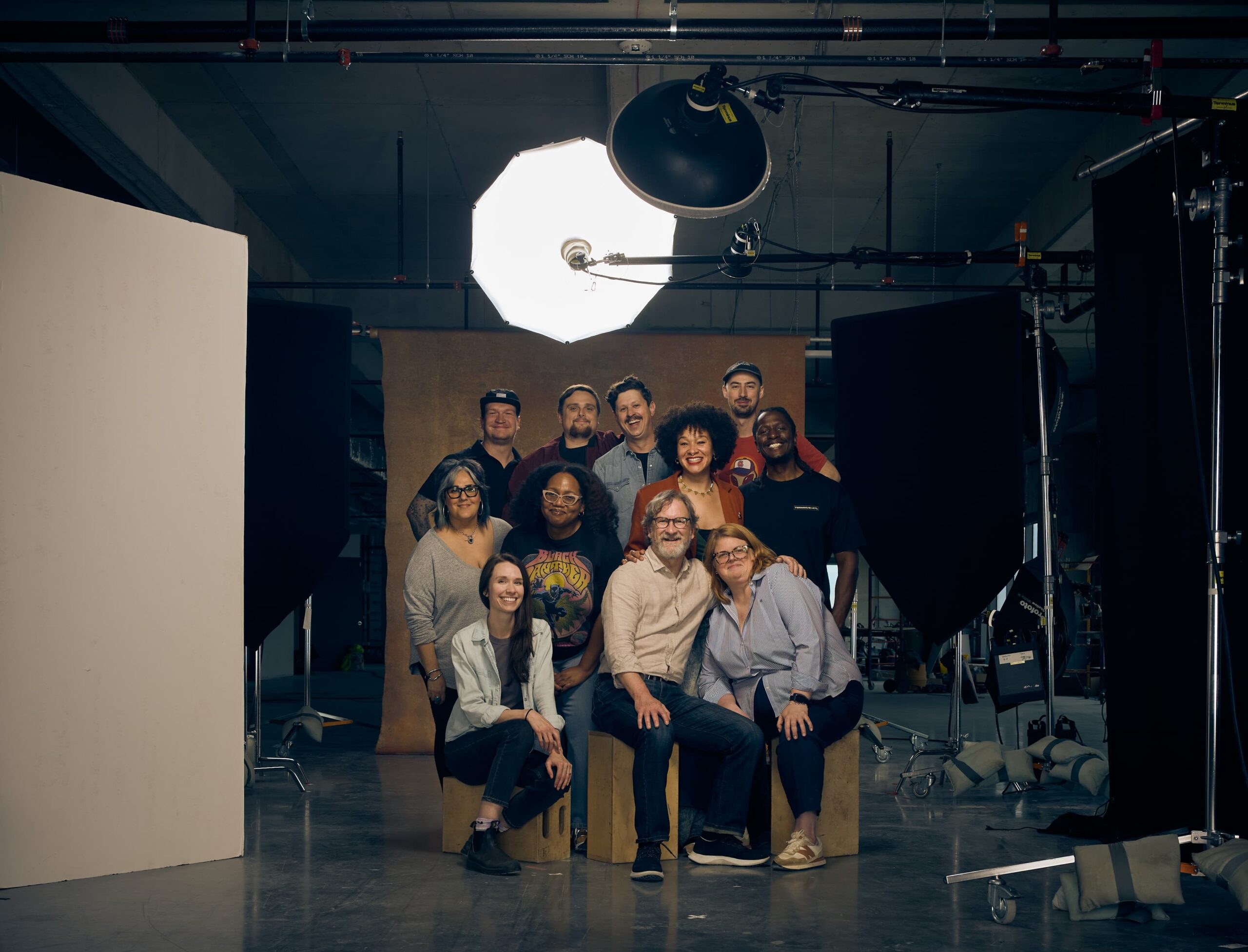 A group of eleven people pose and smile together in a professional photography studio, surrounded by lighting equipment and backdrops.