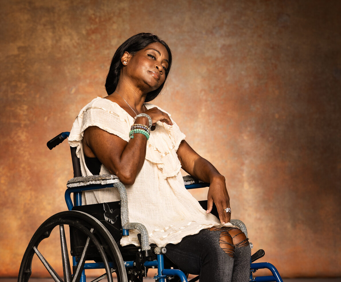 A woman in a wheelchair smiles at the camera. She is wearing a light-colored blouse and black pants, with her hair styled neatly and bracelets on her wrist, sitting against a warm, textured background.
