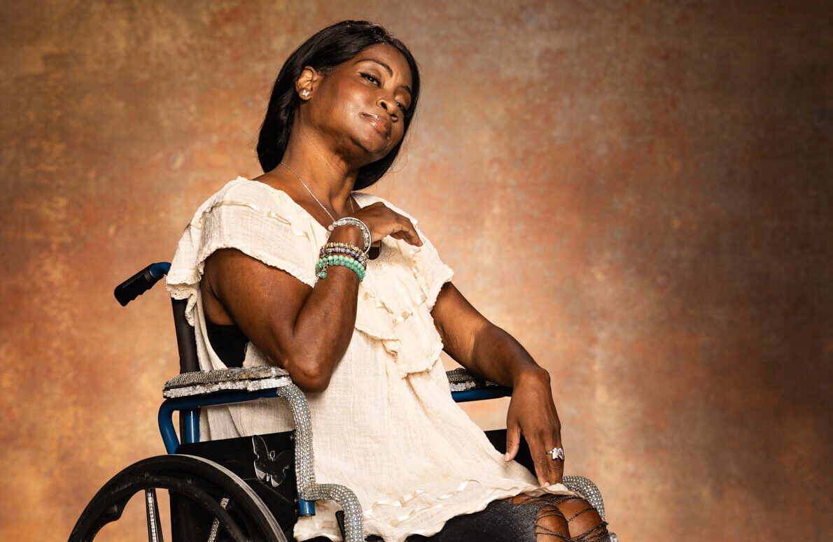 A woman in a wheelchair smiles at the camera. She is wearing a light-colored blouse and black pants, with her hair styled neatly and bracelets on her wrist, sitting against a warm, textured background.