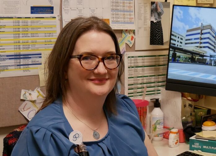A woman with glasses and a blue blouse sits at a desk in an office, smiling. She has an ID badge and is surrounded by papers, a computer monitor, and office supplies.
