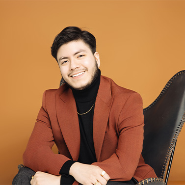 A person with short dark hair and a beard, wearing a rust-colored blazer and black turtleneck, sits smiling on a dark chair against a solid orange background.