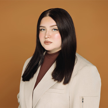 A woman with long straight dark hair, wearing a beige blazer over a brown turtleneck, poses against a solid tan background. She has a neutral expression and natural makeup.