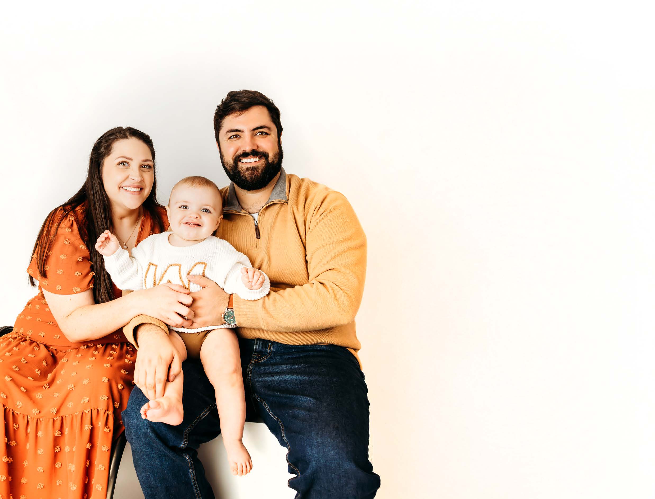 A smiling woman in an orange dress, a man in a tan sweater, and a baby in a white sweater sit together against a plain white background.