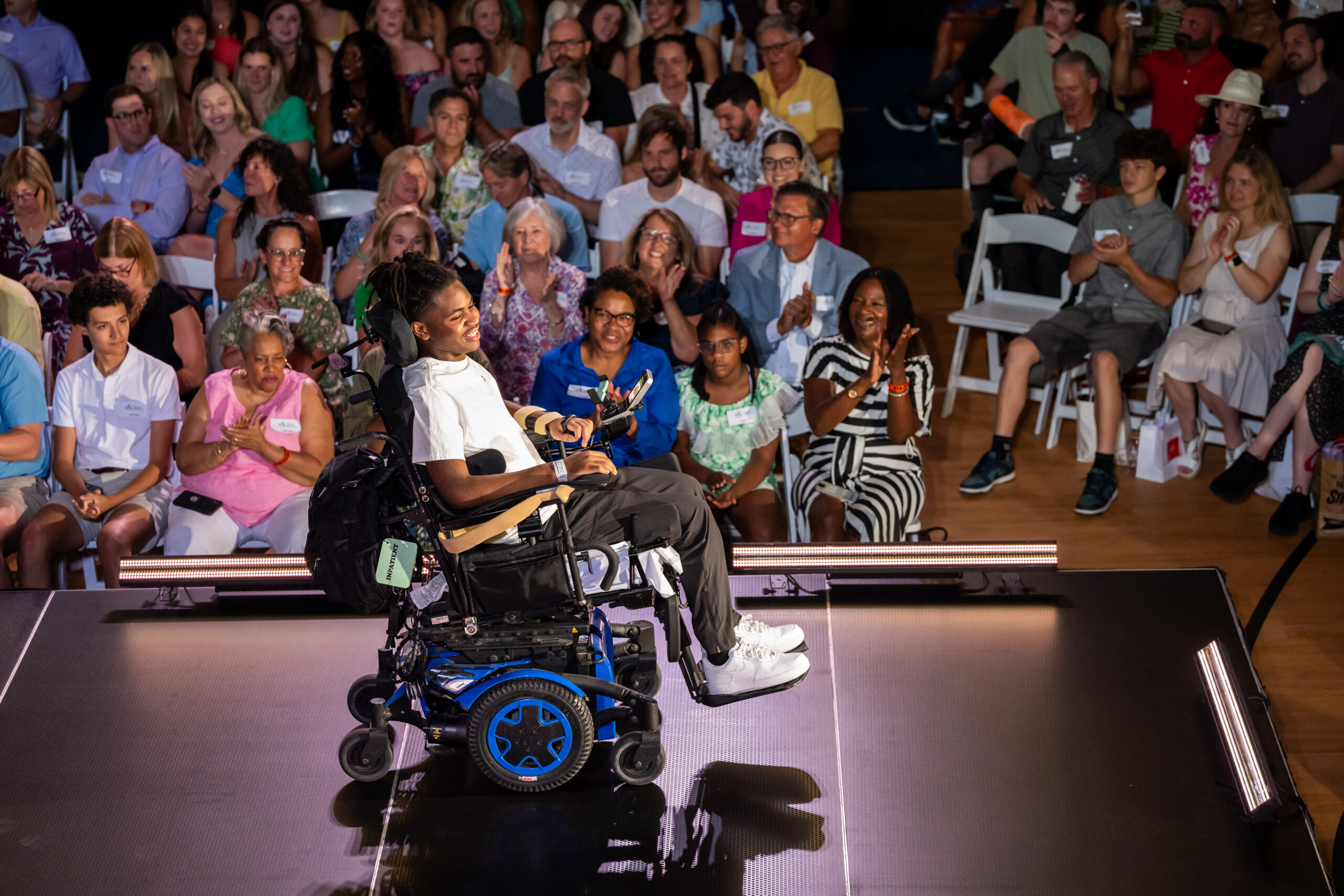 A person in a wheelchair performs onstage under bright lights. The audience, seated and clapping, shows support and attention. The performer is centered, surrounded by diverse spectators, creating an inclusive and energetic atmosphere.