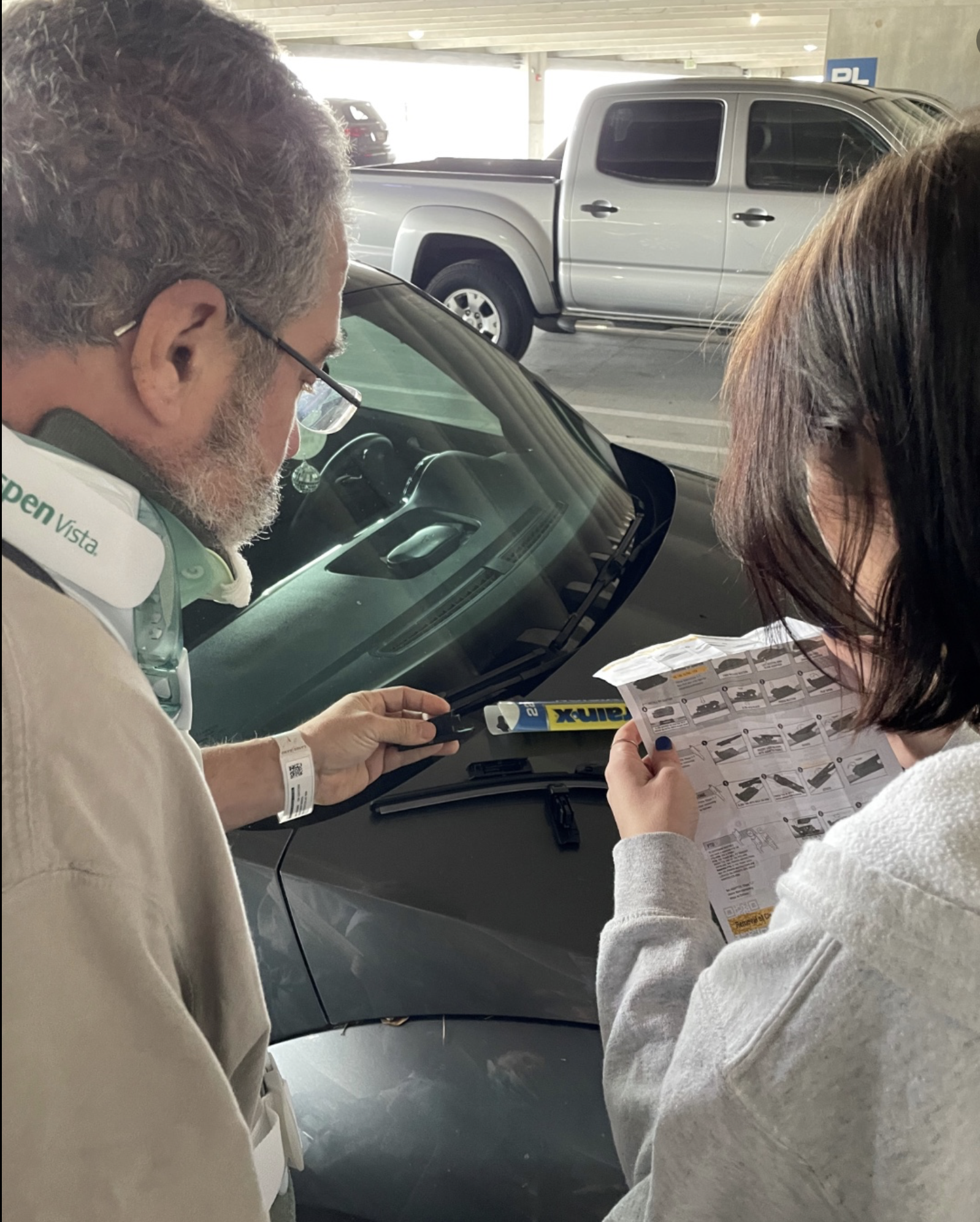 A man and woman stand by a car in a parking garage, part of the day program at Shepherd Center. The man, wearing a neck brace and hospital wristband, holds a wiper blade as the woman reads instructions.