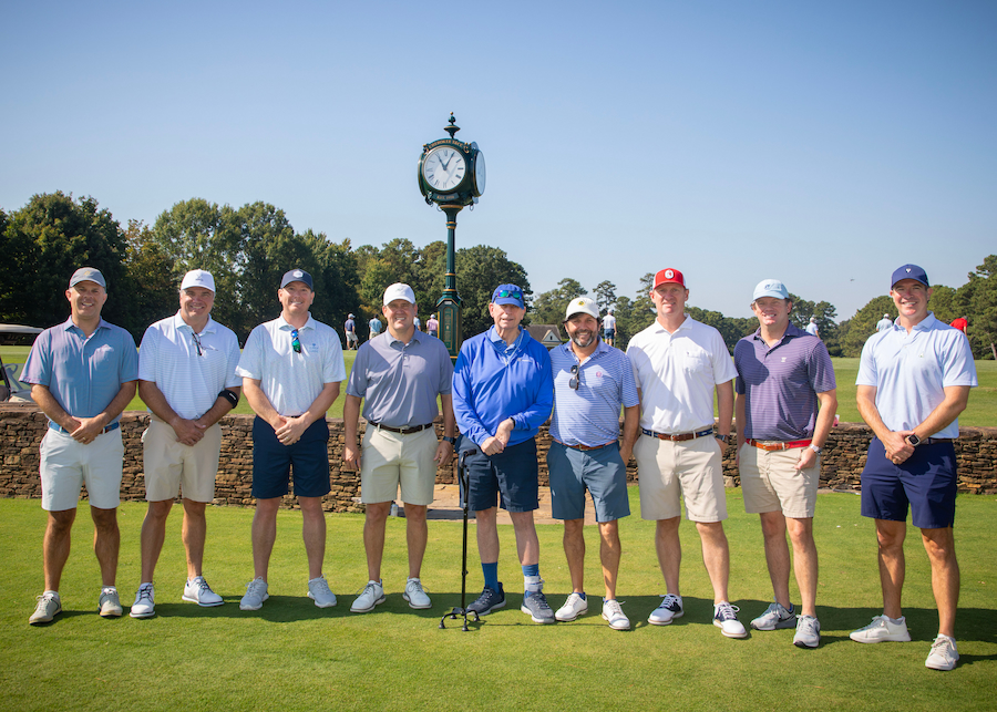 Nine men in golf attire standing in a row on a golf course in front of a tall, decorative clock.