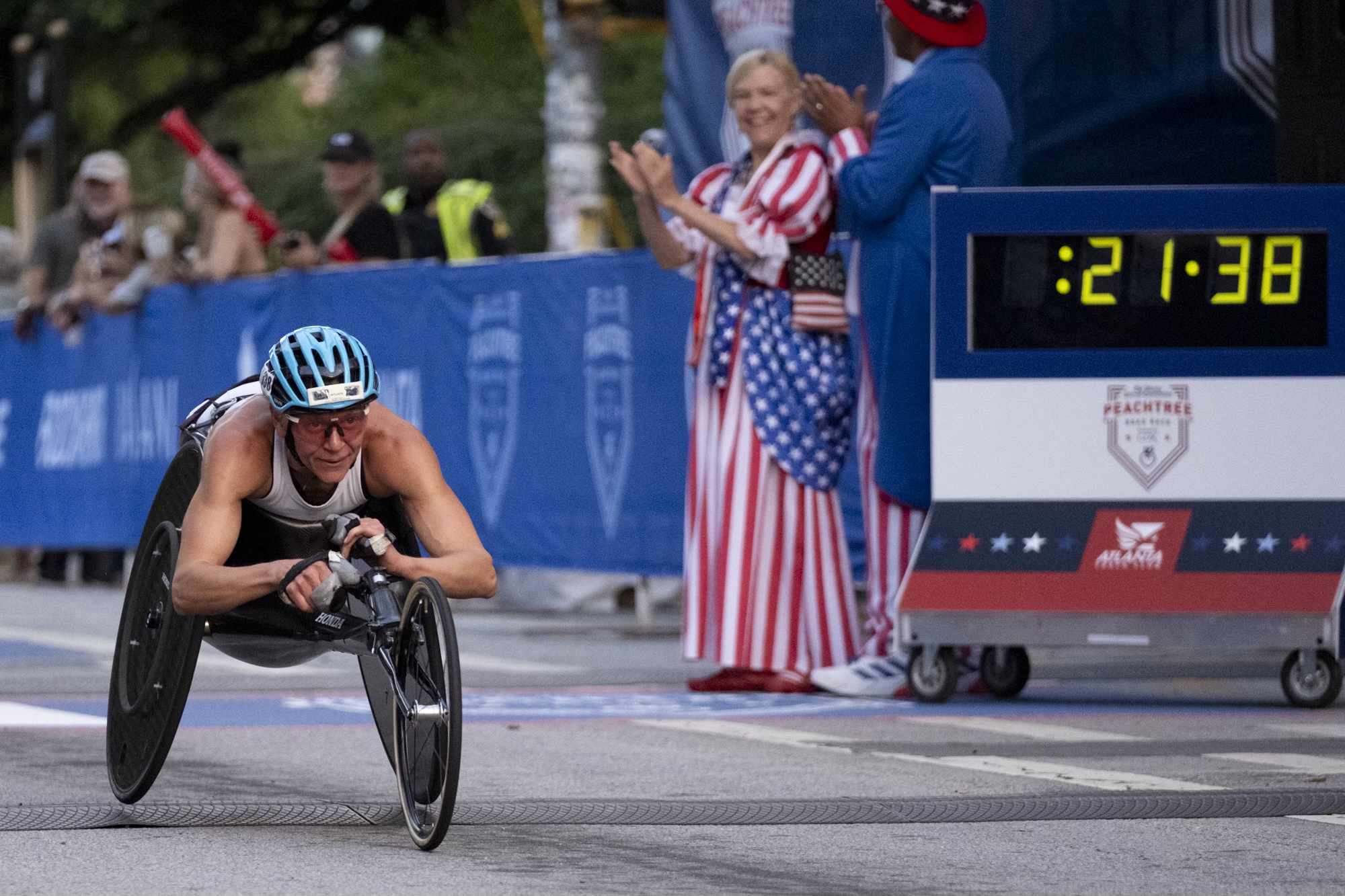 A wheelchair racer crosses the finish line as a digital timer shows 21:38. Spectators cheer by the side of the race.