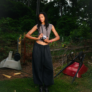 A person with long, dark hair stands outdoors in a garden, wearing a sleeveless top and loose black pants. They hold a camera and are surrounded by gardening tools and equipment with a forested background.