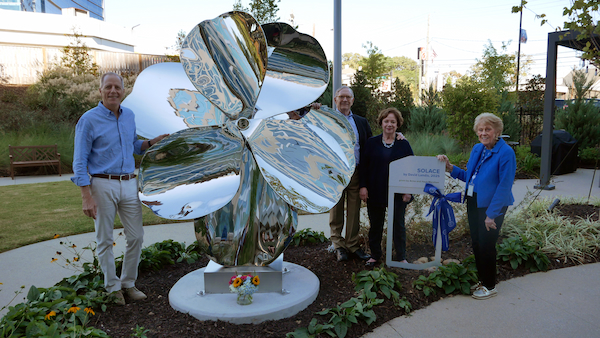 Four people stand around a large, shiny metallic flower sculpture in a garden. One person holds a plaque with a blue ribbon. There are flowers at the base of the sculpture and greenery surrounding them.