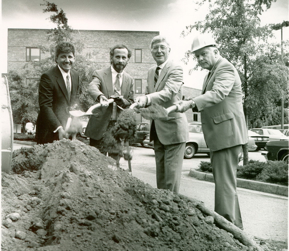 Four men in suits and a hard hat participate in a ceremonial groundbreaking, smiling while shoveling dirt. A building and parked cars are visible in the background.
