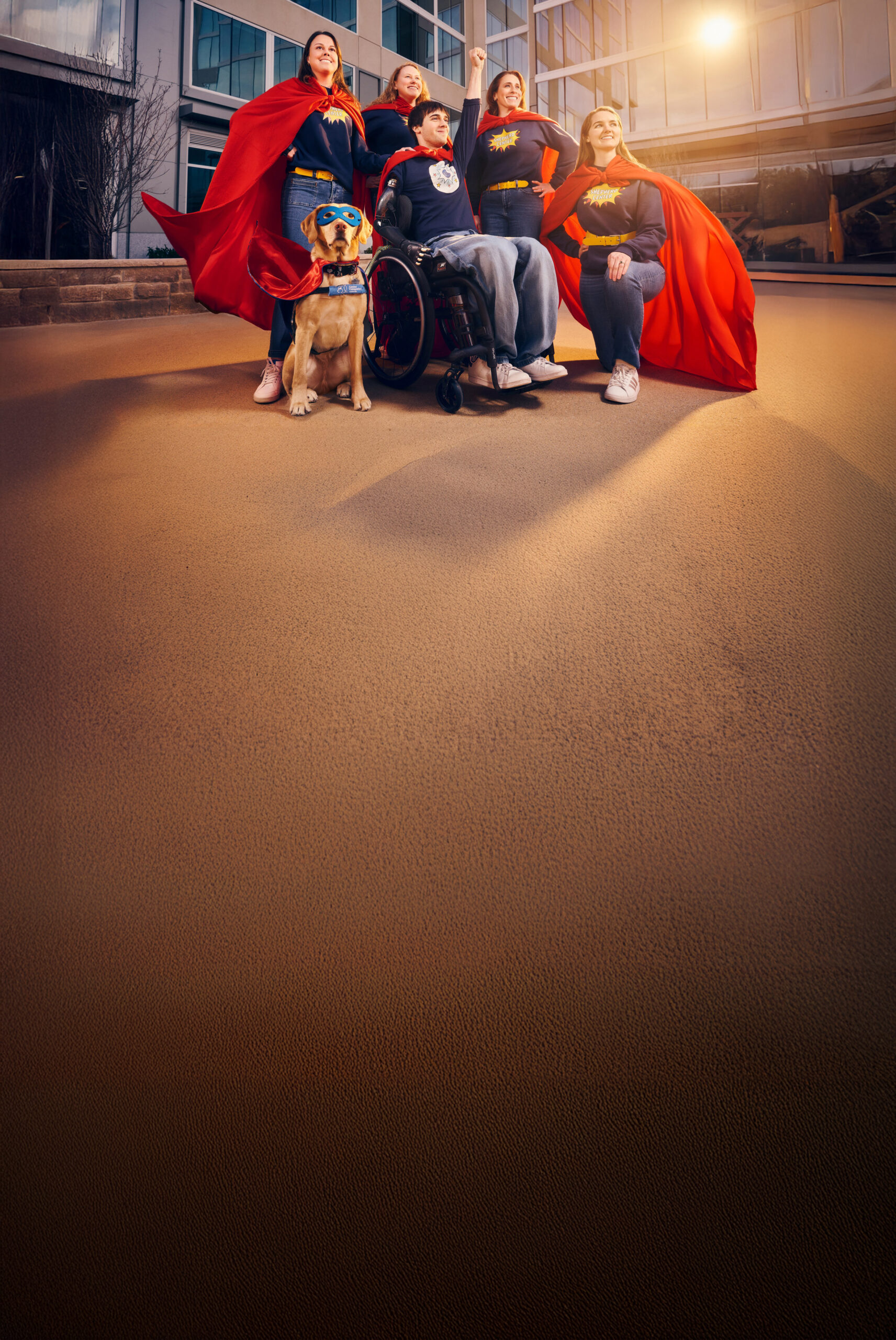 Five people wearing superhero costumes and capes, including one person in a wheelchair, pose triumphantly outdoors. A dog wearing a superhero costume sits in front. A modern building is in the background.