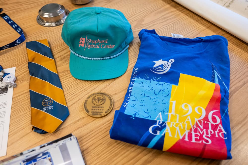 A colorful 1996 Atlanta Paralympic Games t-shirt, blue-and-yellow striped tie, turquoise Shepherd Spinal Center cap, medal, and lanyard are arranged on a wooden table.