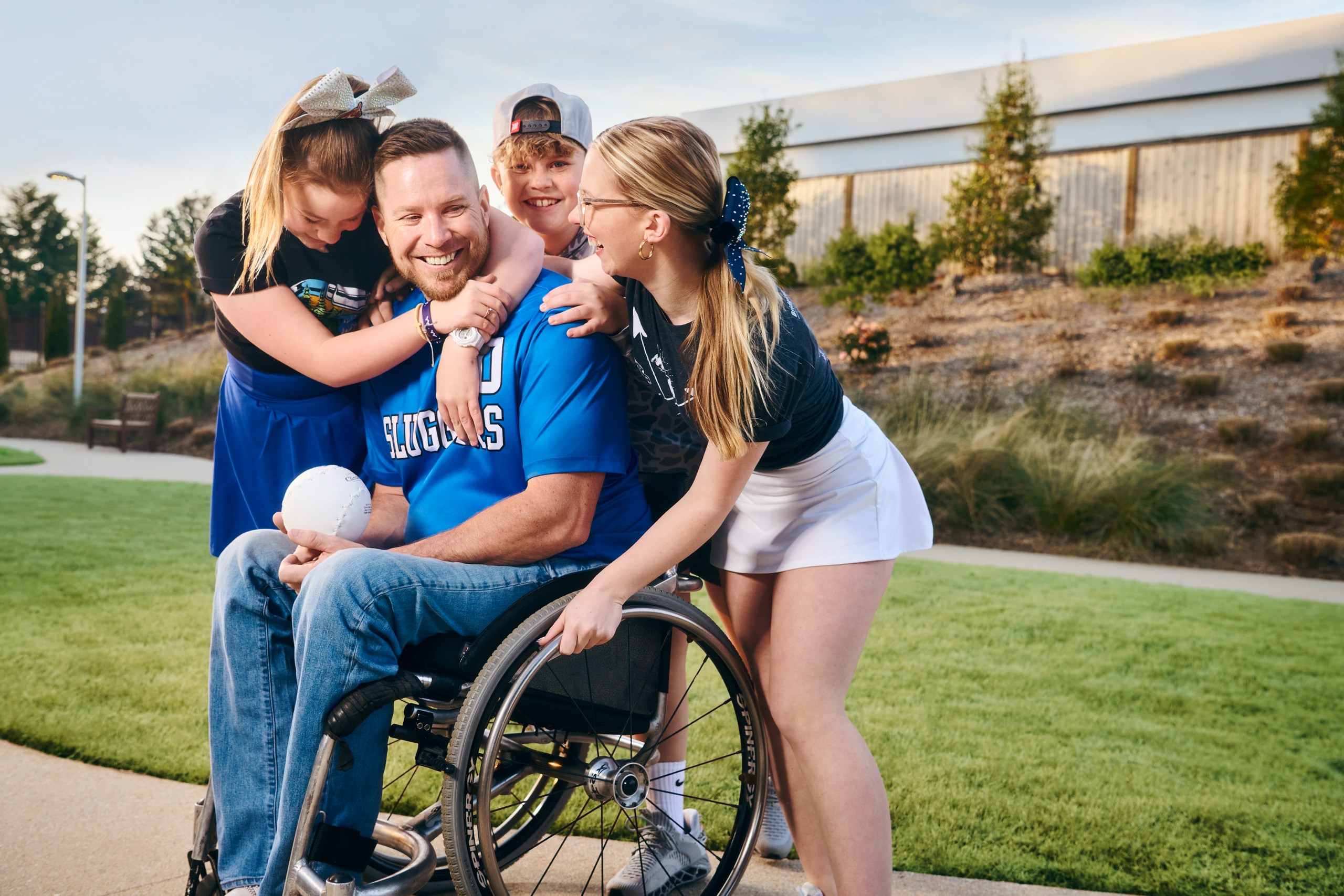 A smiling man in a wheelchair holds a ball while three happy children hug him outdoors on a sunny day, all wearing sports or casual clothes, with green grass and trees in the background.