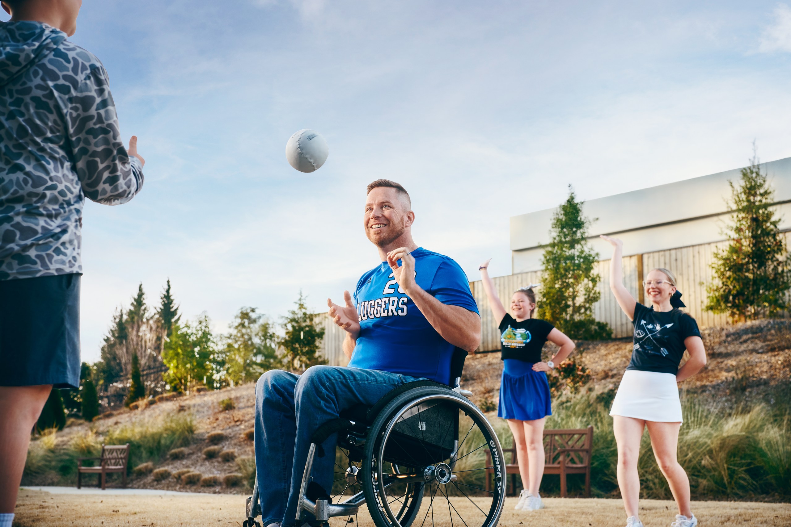 A man in a wheelchair smiles and tosses a football to a child, while two cheering teens watch and laugh outdoors on a sunny day.