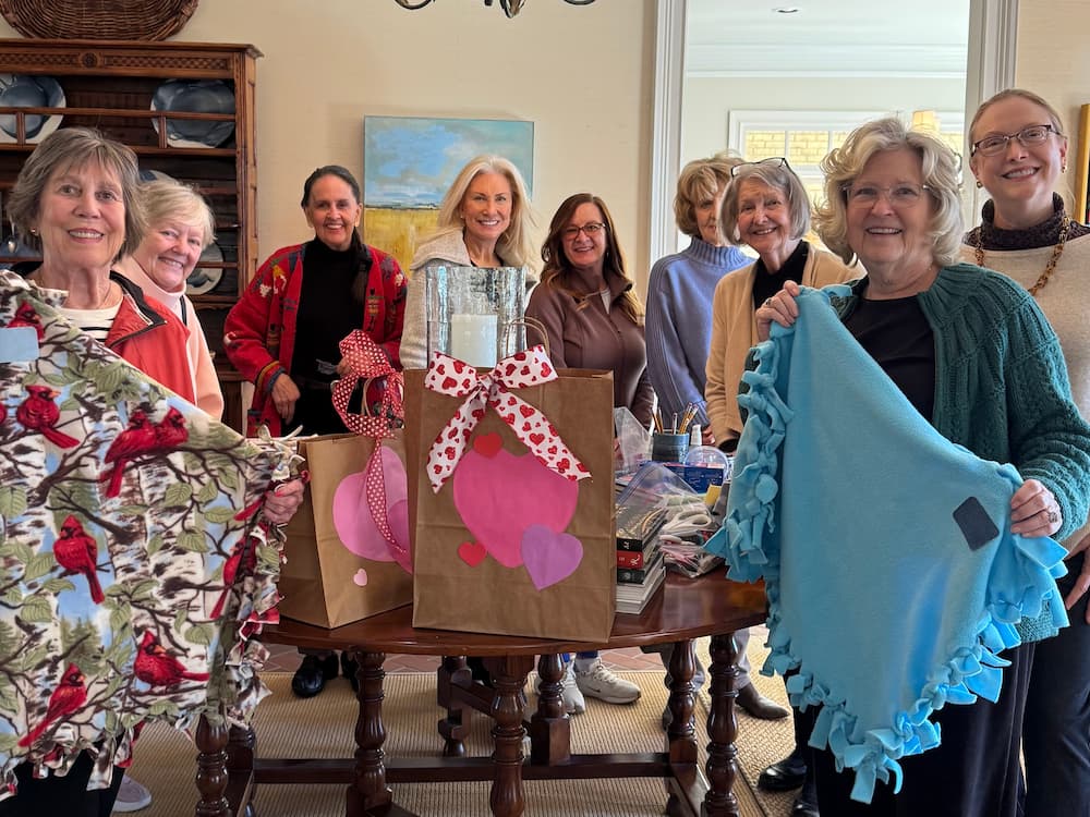 A group of smiling women stands around a table with decorated gift bags. Two women hold handmade fleece blankets, one with a cardinal print and one blue.