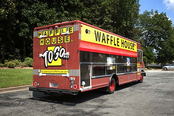 waffle-house-food-truck A red and yellow Waffle House To Go food truck is parked on a paved lot, surrounded by trees and greenery. The truck has large Waffle House signs and service windows on its side.