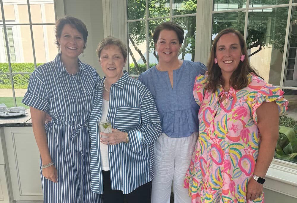 Four women stand together indoors by large windows, smiling at the camera. Three wear brightly patterned or striped dresses and one wears a blue shirt with white pants. Lush greenery is visible outside the windows.