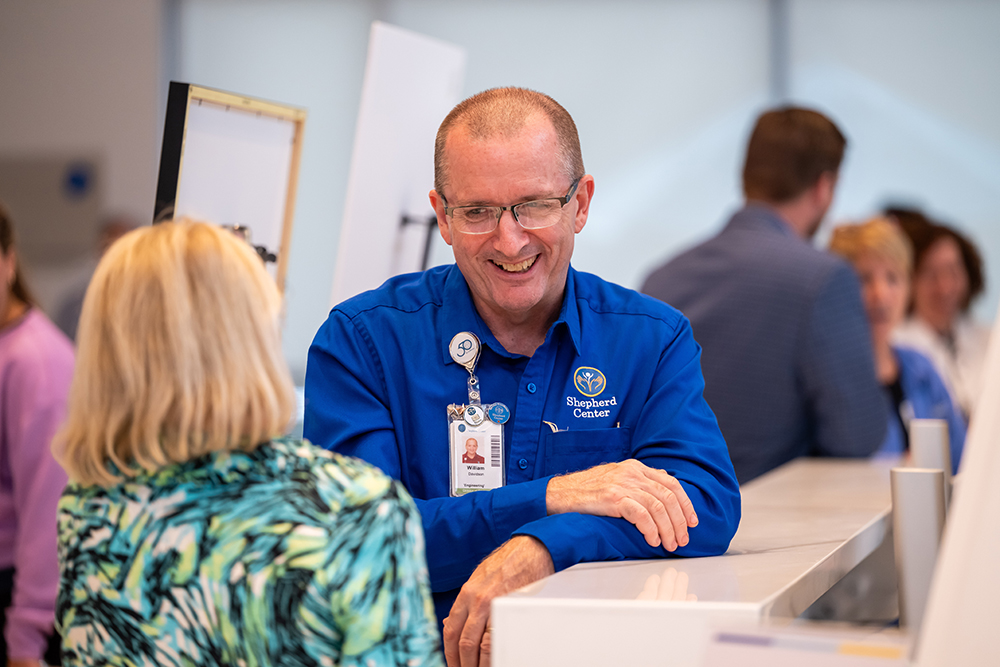 A man in a blue shirt with a name badge smiles while talking to a woman across a counter in a bright, busy indoor setting with several people in the background.