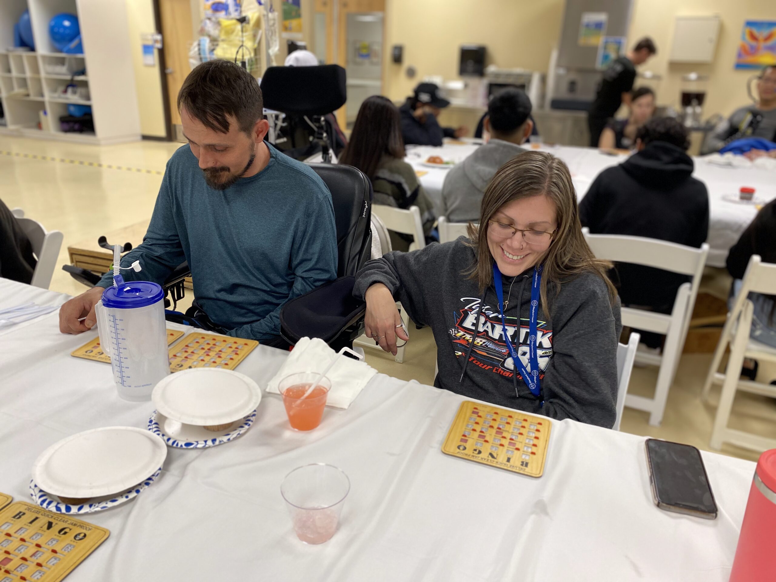 A man and a woman sitting at a table playing bingo. The woman is smiling and holding a bingo card. There are paper plates, a large water bottle, and drinks on the table. Several people are in the background.